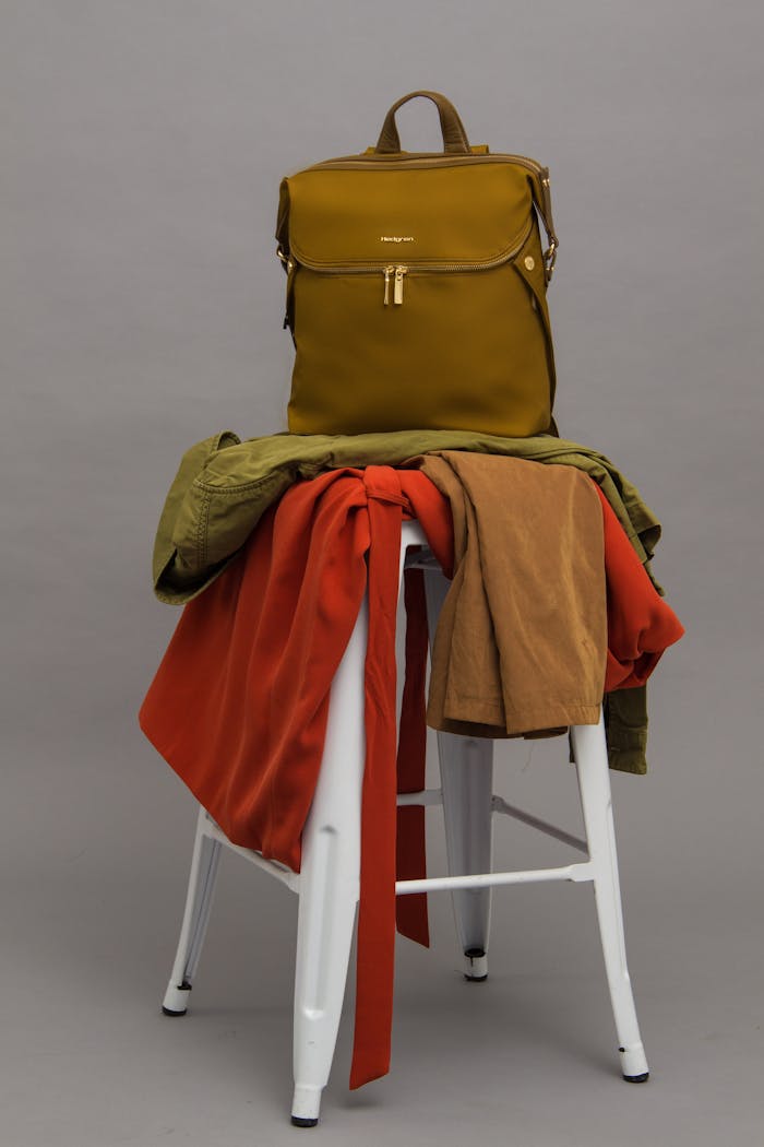 Mastering the First Impression: Your intriguing post title goes here A fashionable olive green backpack placed on a white stool with vibrant clothes draped over it.